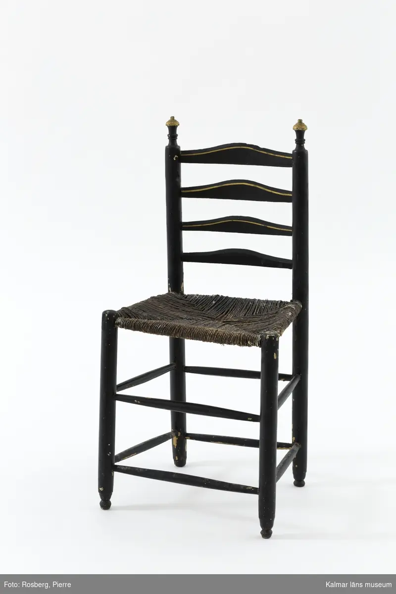 KLM 18989:3. Stol, stegstol, stegryggstol. Av typen Ladderback chair - stegryggstol. Av trä. Ryggstöd med fyra bladformade och tvärgående ryggbrickor. Ryggstolparna avslutas med snidade detaljer. Raka ben med dubbla rader av runtgående benslåar. Målad i svart med detaljer i guld. På ett flertal ställen syns att stolen varit målad i ljust gul, troligen originalfärg. Flätad sits av rotting i tre lager.