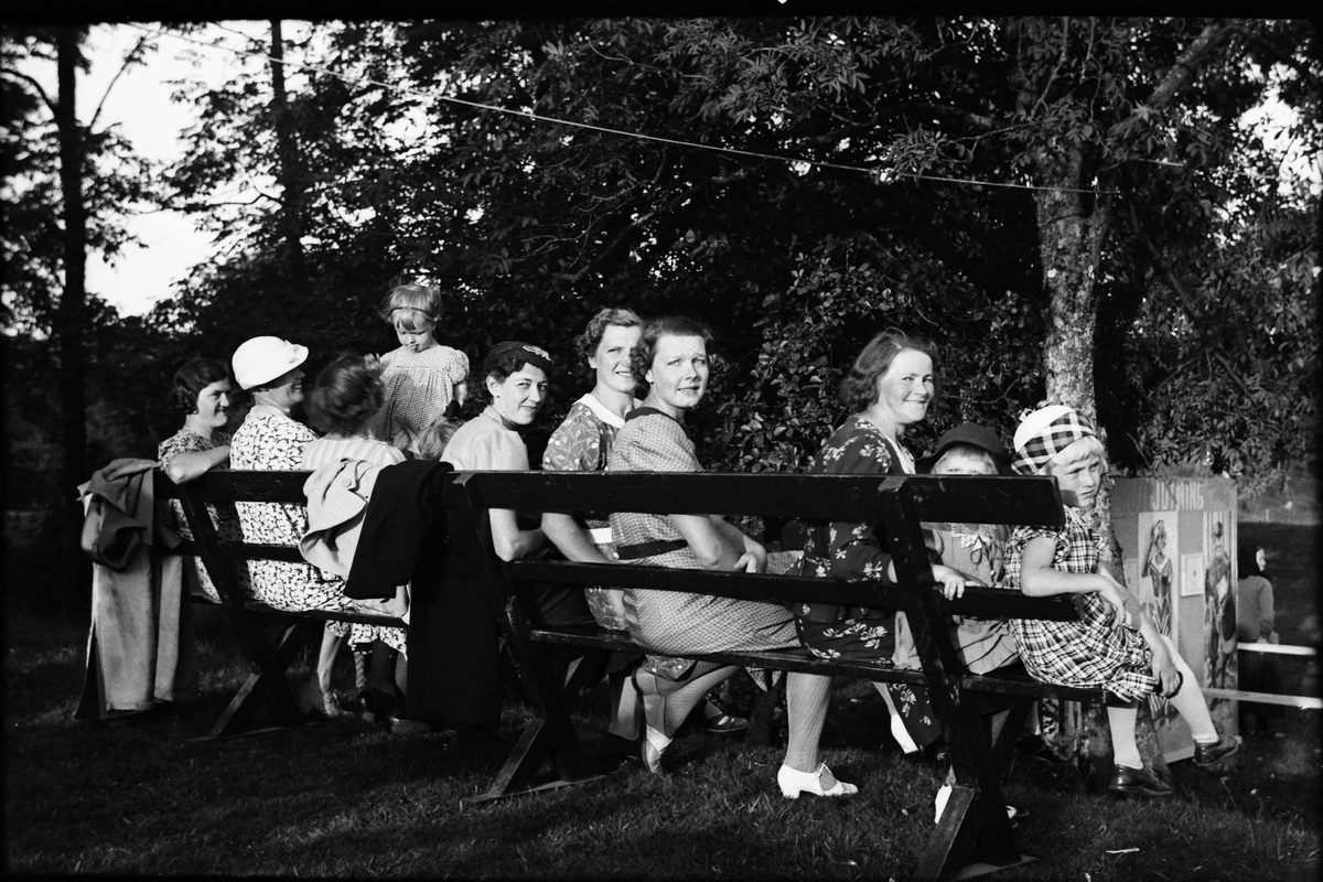 Kvinnor och barn sitter på en bänk i en träddunge på en höjd över en prickskyttebana på en festplats.