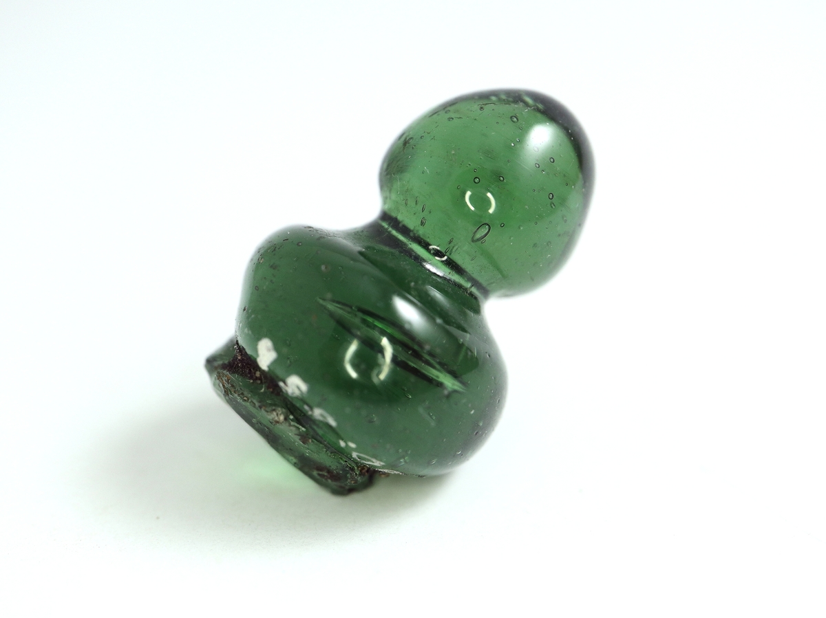 Den översta profilerade knoppen av ett smaragdgrönt glaslock. Ojämn i formen. Fullt av jämnt fördelade blåsor i glasmassan. Möjligen till en pokal eller ett Hertig Carlglas med lock.