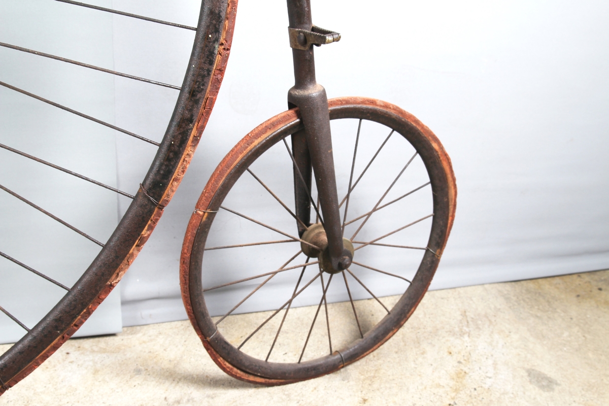 Svartlakkert velosiped med stort forhjul og lite bakhjul. Pedalene er montert direkte på fornavet og setet er plassert rett over.