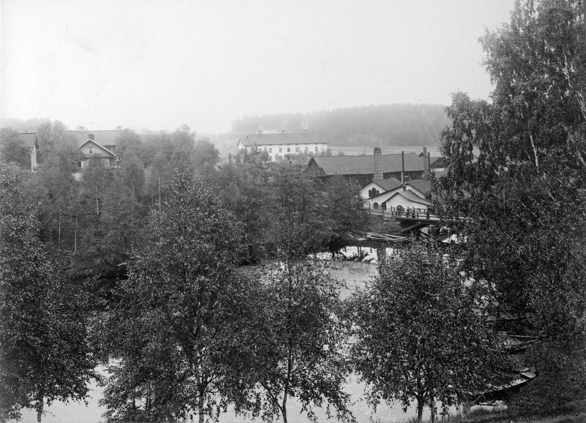 Kolsva Jernverk sedd över ån. På bron står några personer och tittar på en man som metar. Omkring 1900.