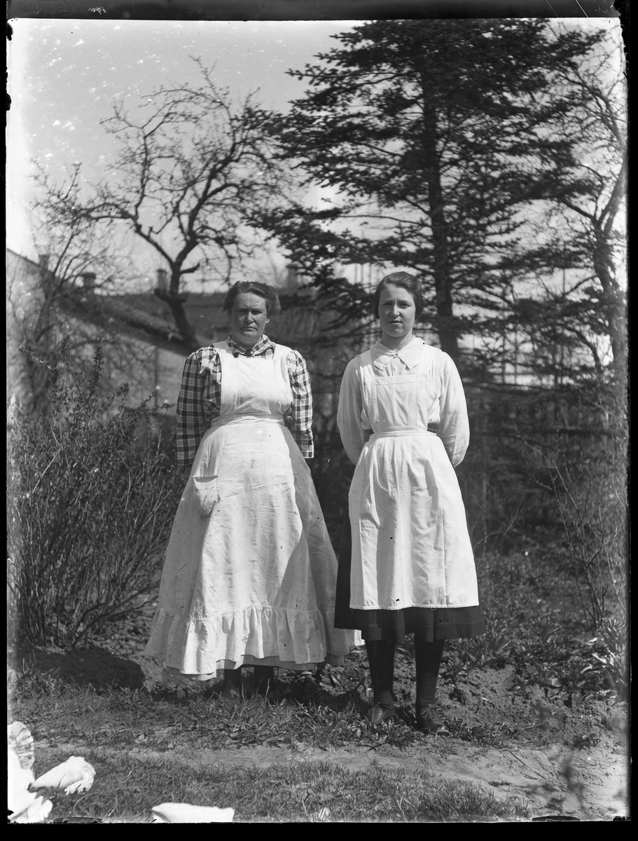 Anna Glasin och Hedvig iklädda förkläden och med händerna på ryggen fotograferade utomhus. I bakgrunden skymtar byggnader mellan träden.