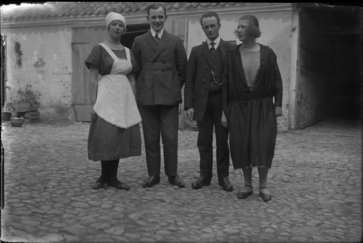 Två kvinnor står på en kullerstensklädd innergård, emellan dem står två kostymklädda män. I fotografens egna anteckningar står: Danmarksresan, Anna Hansén med kamrater.