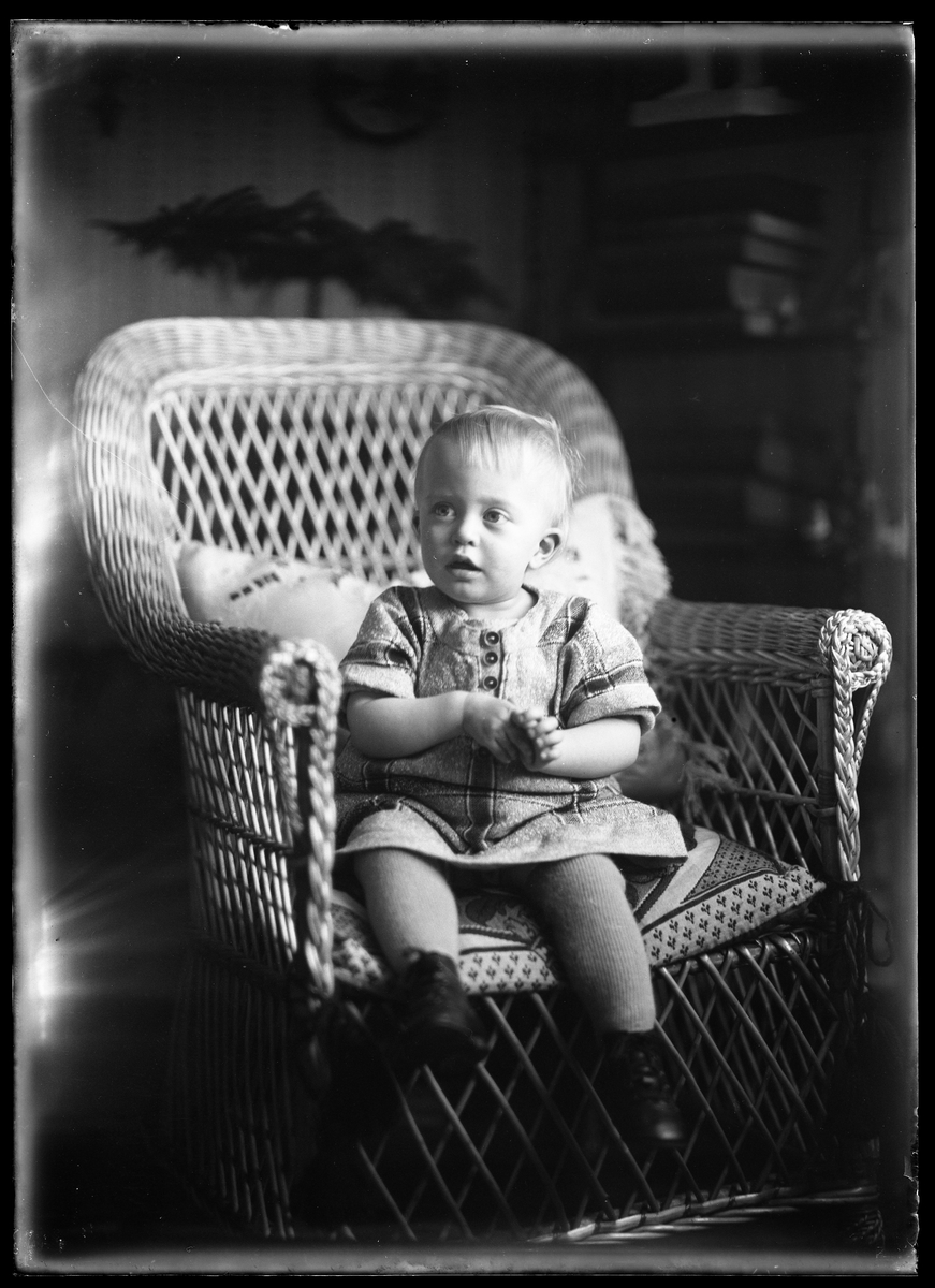 En liten flicka i rutig klänning och svarta skor sitter i en korgstol. I fotografens egna anteckningar står det "Richard Bergs flicka".