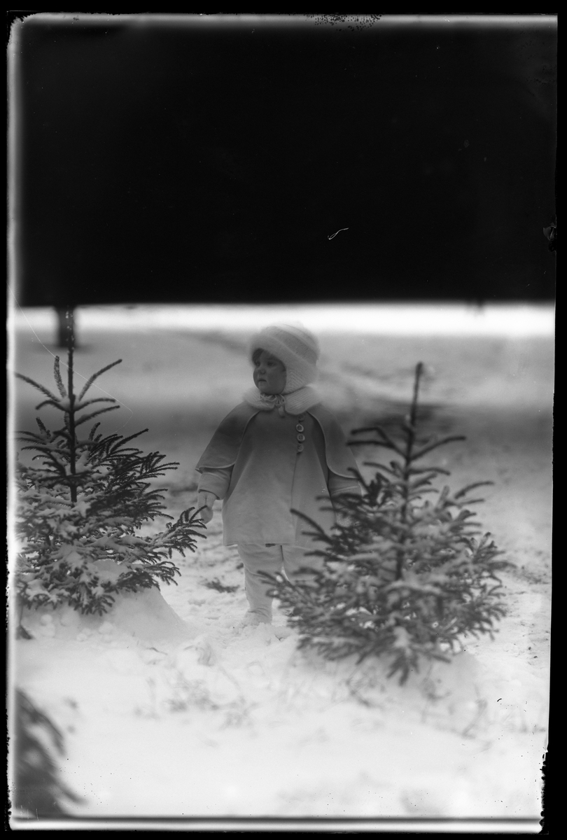 En liten flicka, tolkad som Elsa Margareta Berg, i vita vinterkläder står i snön mellan två små granar. I fotografens anteckningar står det "Ing[enjör] Bergs flicka". Bilden är inte helt exponerad.
