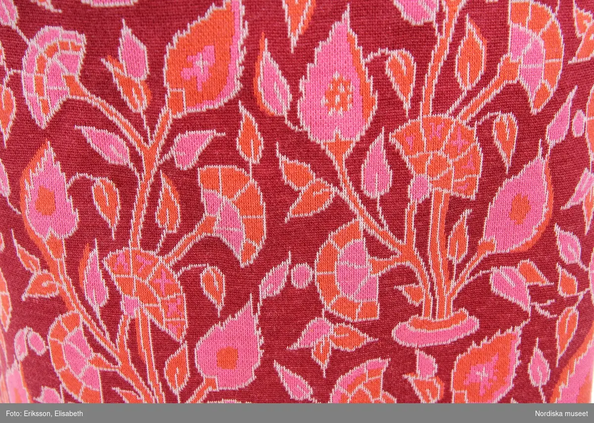 Dräkt, jacka och kjol, i bomullstrikå med tryckt mönster nejlikor i rosa, orange och rött. Jackan, fyrkantig modell, med rundringad hals och knäppning fram, bandkantad rosa och röd jersey. Kjolen a-line modell, knäkort, med dragkedja i sidan. Etikett i nacken, storlek 42. / Helen Persson, 2018-07-13