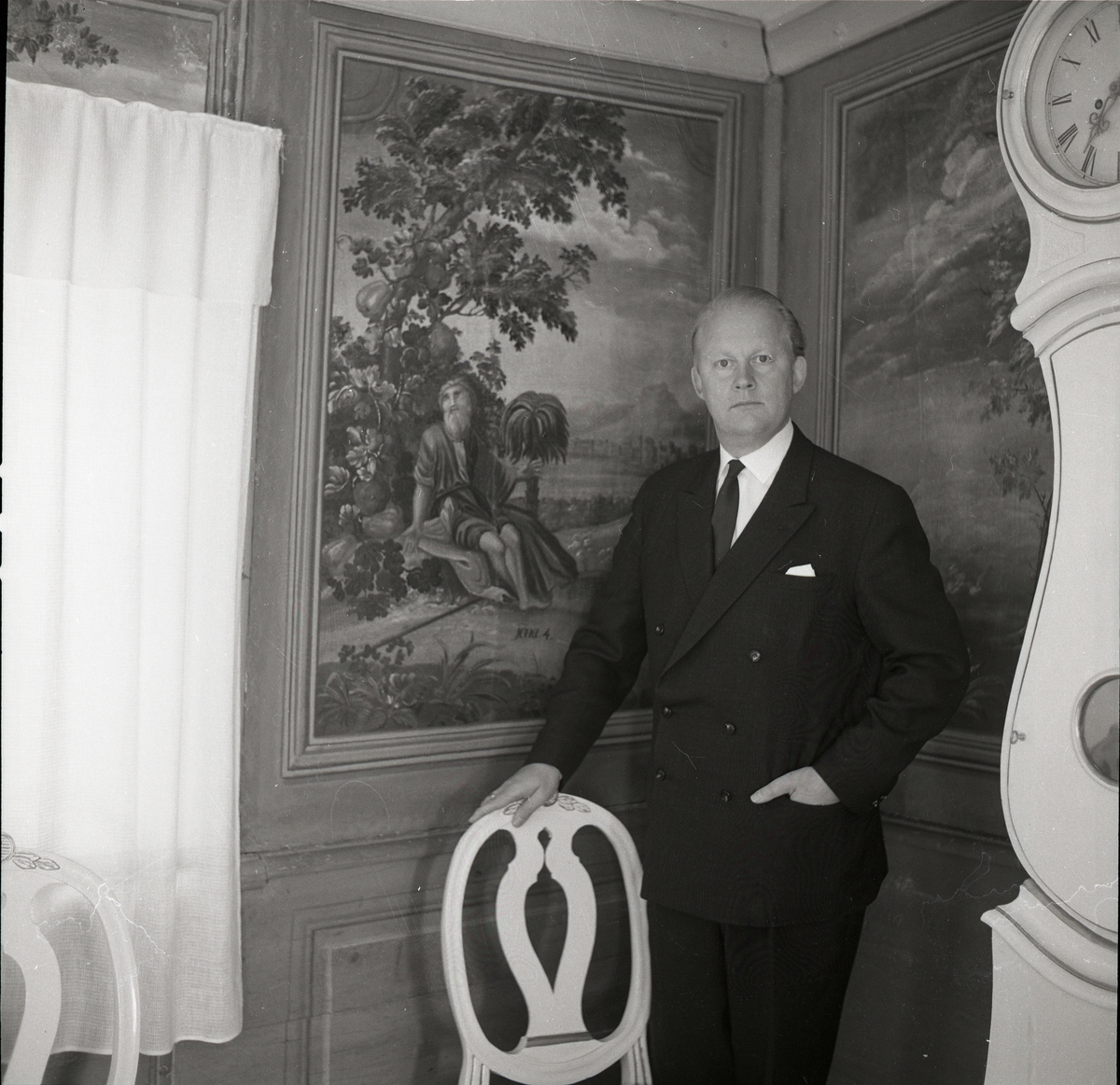 Författaren och diktaren Bo Setterlind står i en interiör med väggmåleri. Ena handen vilar på en stol och bredvid står ett golvur, 26 juni 1968.