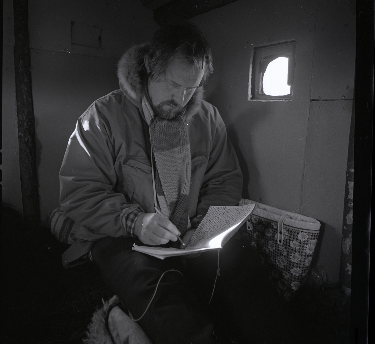 En man iförd jacka och halsduk sitter i en koja på Stråsjöberget och skriver i en bok, 1973.