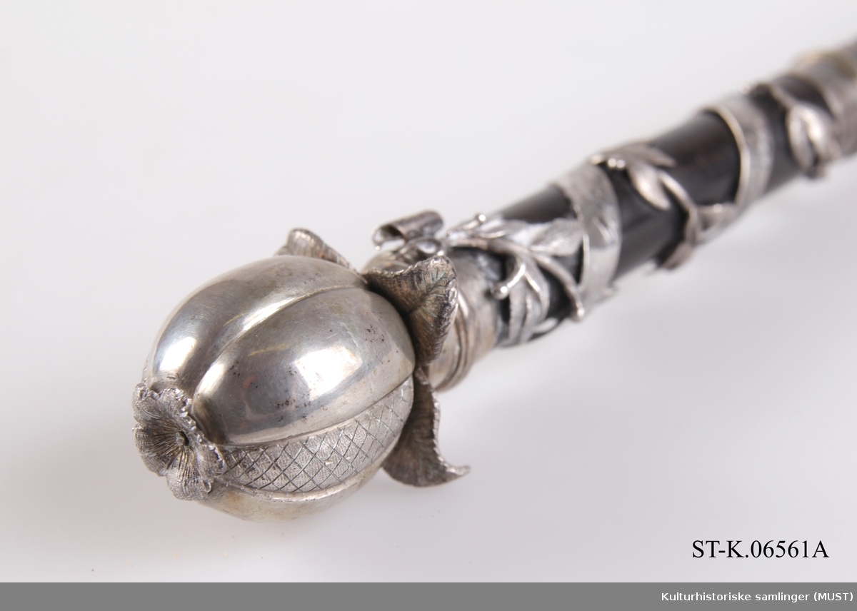 Dirigentstav dekorert med et granateple i sølv på toppen. Håndtaket er omviklet med en lauvbærranke og et innskriftsbånd i sølv.