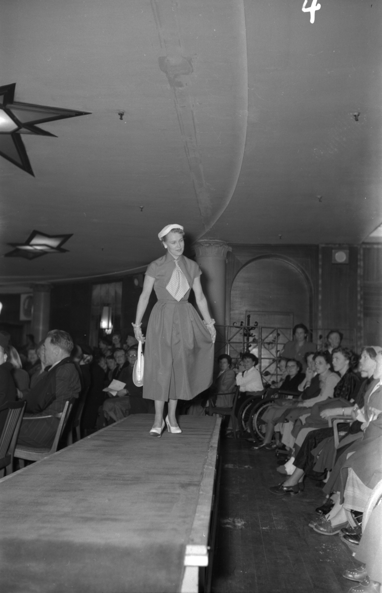En kvinne på podiet iført kortermet kjole. Fra en motevisning på Steen & Strøm fra 7. mai 1954..