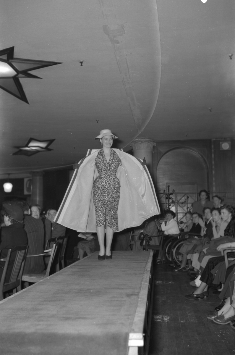 En kvinne på podiet iført mønstrete kjole og kappe. Fra en motevisning på Steen & Strøm fra 7. mai 1954..