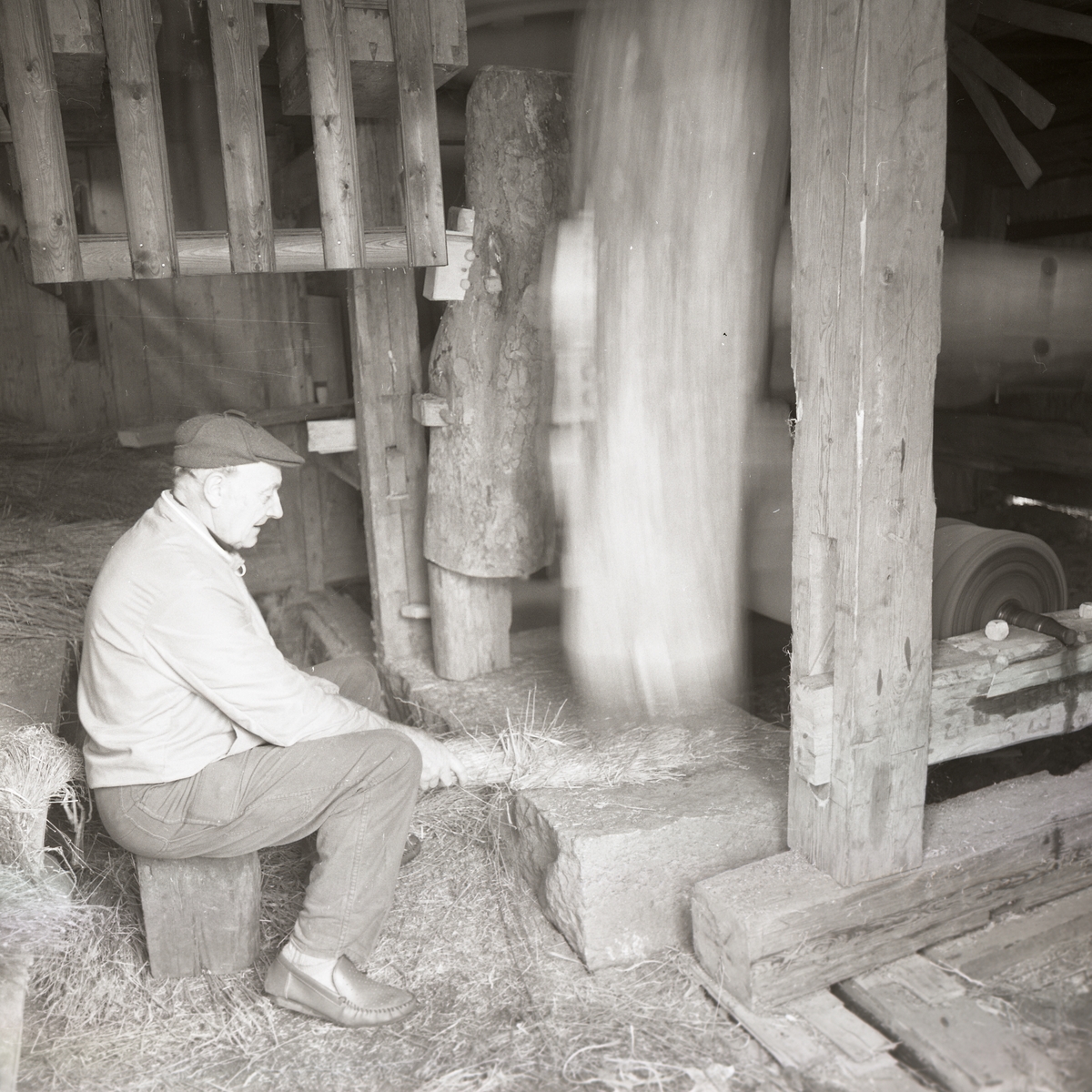 En man sitter med lin vid en linstamp gjord av träplankor, trästolpar och sten, hösten 1974.