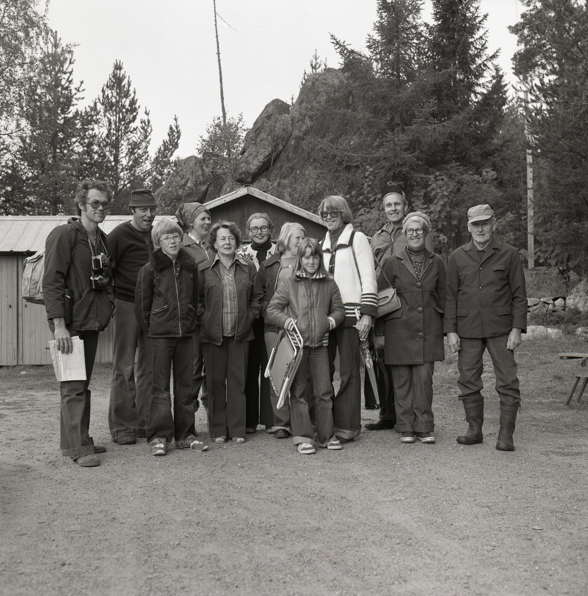 En grupp människor står på en grusväg framför en byggnad och poserar. En flicka håller i en campingstol och en man har en kamera runt halsen, några har glasögon och några har huvudbonader. Bilden är tagen i samband med invigning av en kyrkstig vid Tannåsen, september 1977.