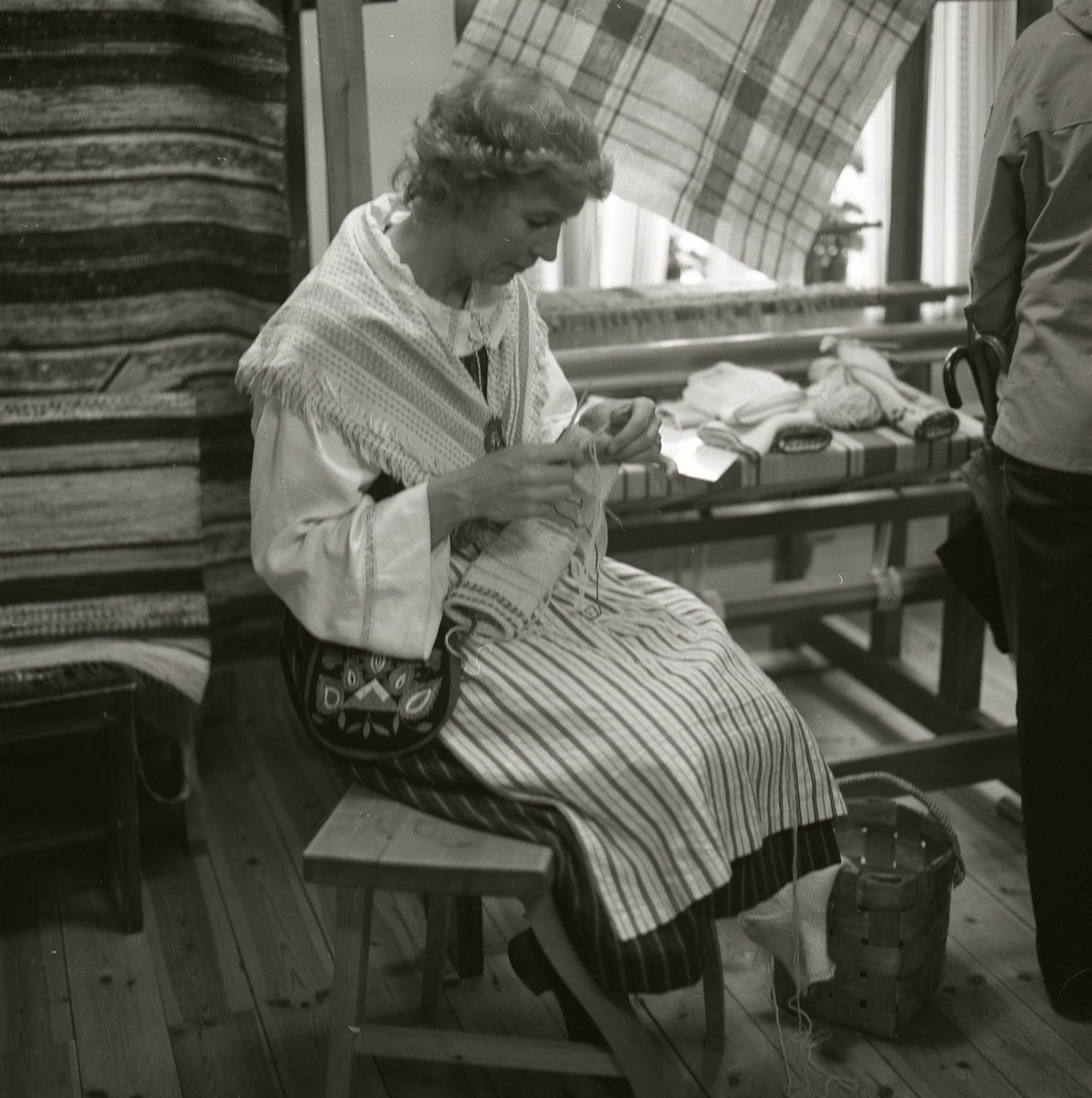 En kvinna iklädd folkdräkt sitter på en pall och fäster trådar på ett vävt föremål under Gammelvärldens dag vid Hembygdsbyn 1980. Till höger om henne står en vävstol och bakom henne hänger vävda mattor.