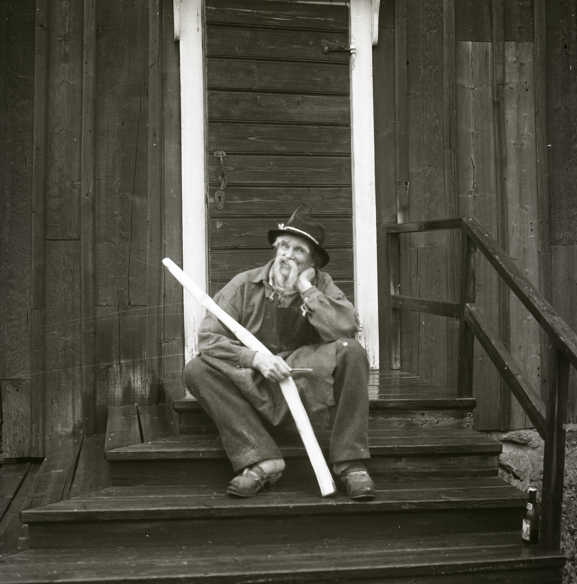 På en trappa till en timrad byggnad sitter en utklädd man med huvudbonad, förkläde, träskor och en lång mustach. Han håller i ett träföremål och en kniv. Bakom honom finns en dörrkarm samt en dörr med handtag, nyckelhål och beslag. Trappan har ett räcke och på det nedersta trappsteget står en glasflaska, Hembygdsbyn 1980.
