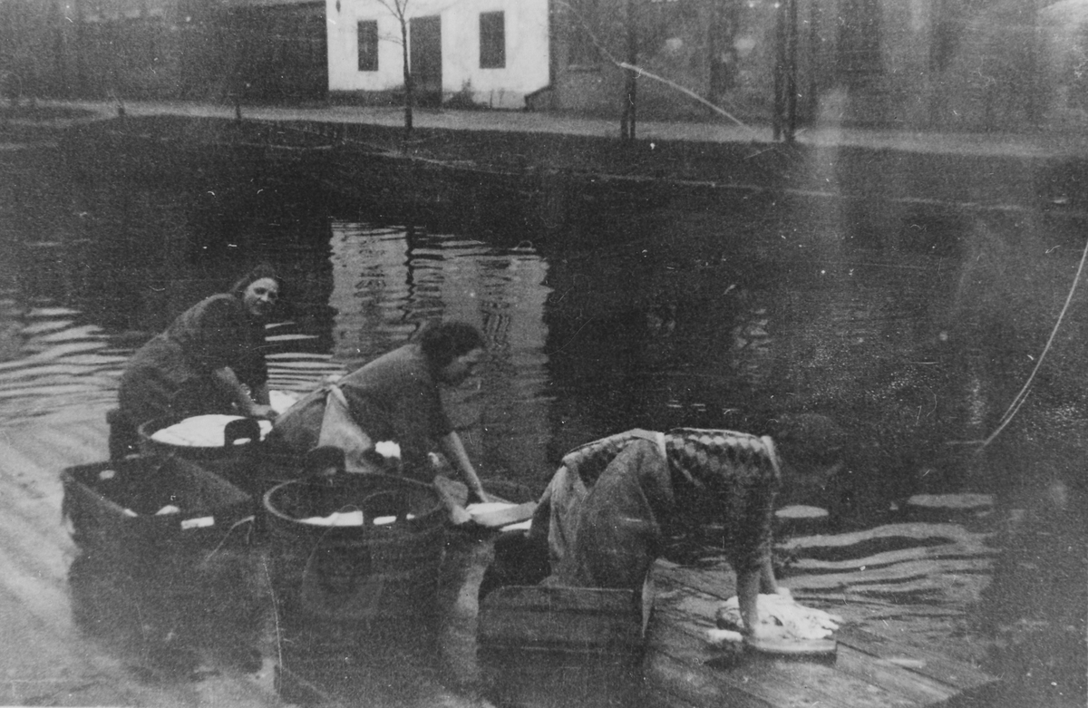 Tre kvinnor står på knä på en tvättbrygga och tvättar tvätt i Lillån.