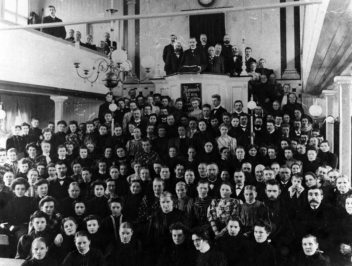 Bilden visar ett 50-tal kvinnor och män som står i missionskyrkans kyrksal. Vissa uppe på läktaren till vänster samt uppe på predikstolen.