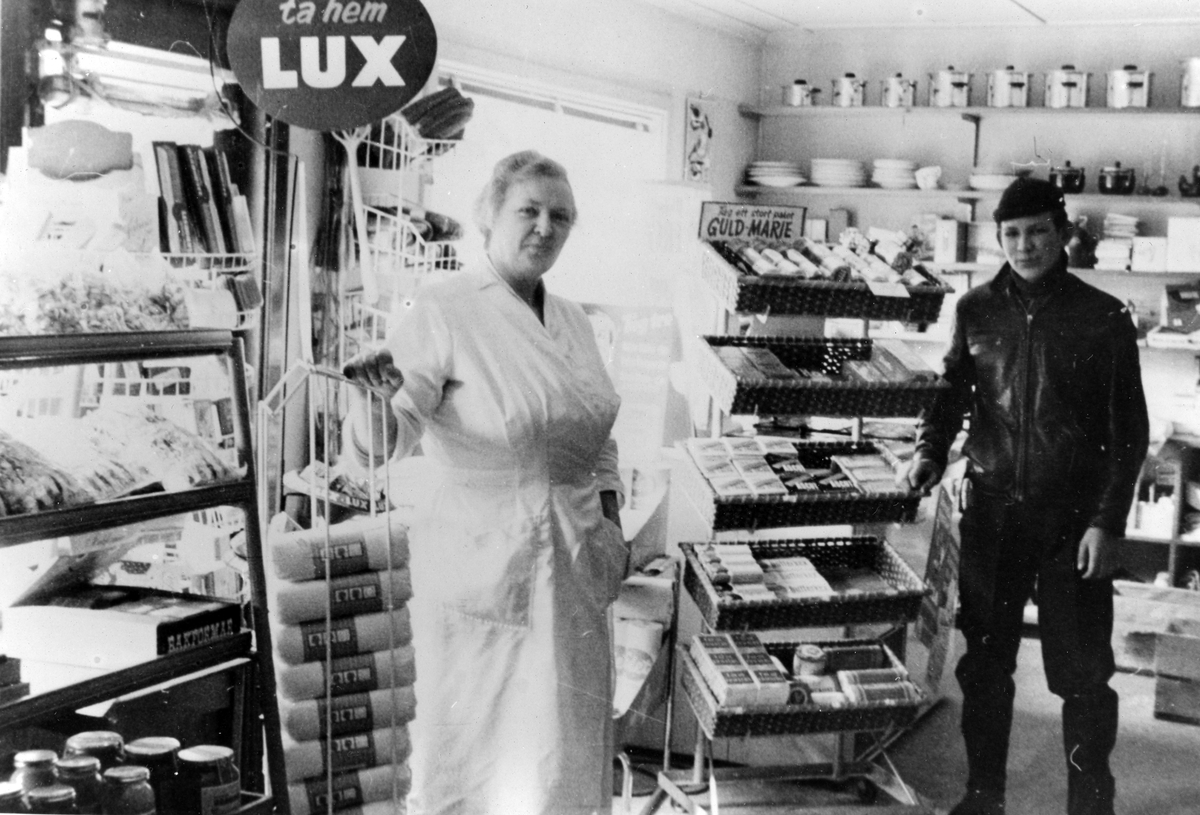 Inifrån Kjells Speceriaffär. Till vänster en kvinna i vita arbetskläder och till höger en man. Reklamskyltar med "Lux" och "Guld Marie".