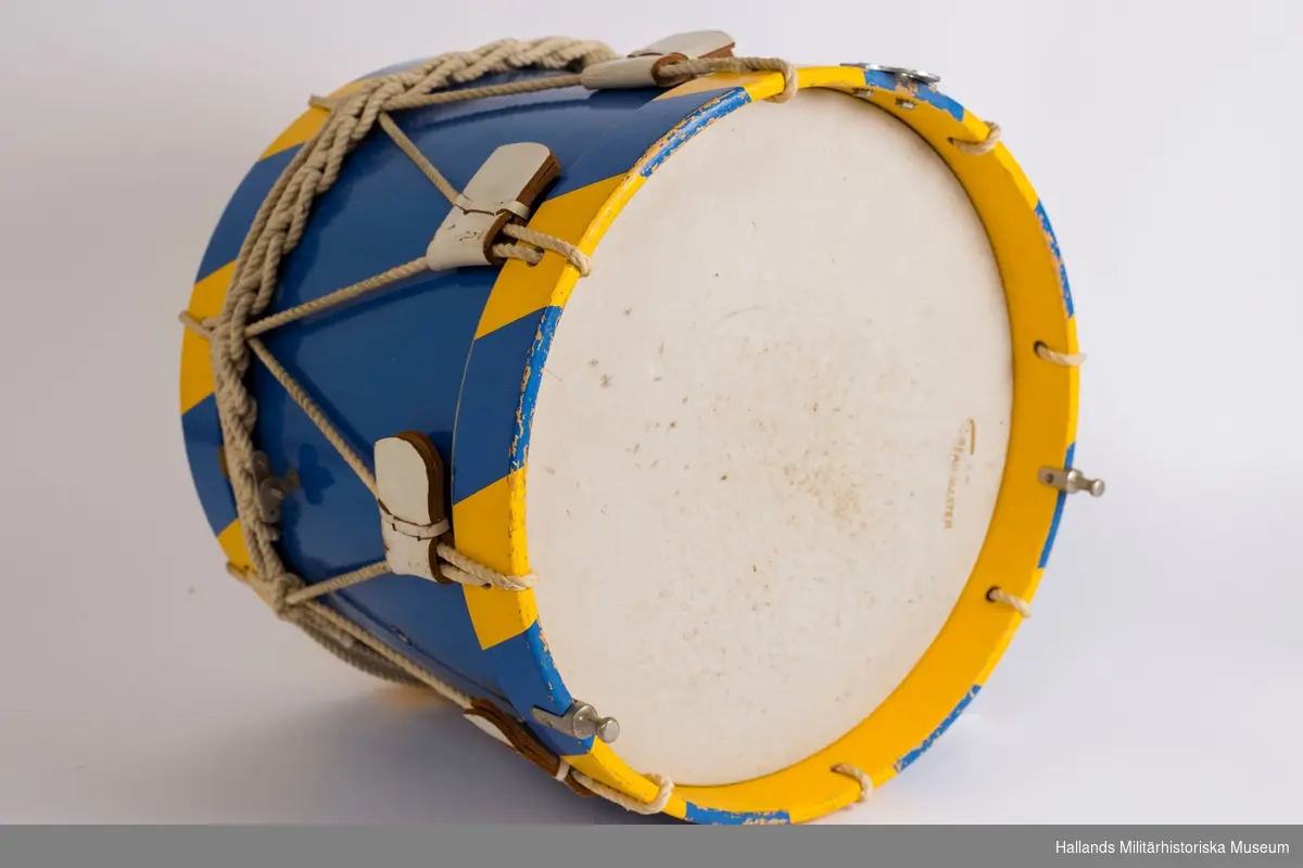 En blå trumma i trä med metalldetaljer. Trummans kanter är blå- och gulrandiga. Vita rep spända mellan båda träramarna, runtom hela trumman och med vita läderdetaljer. Vitt slagskinn. Trumman har ett borrhål på ena sidan.