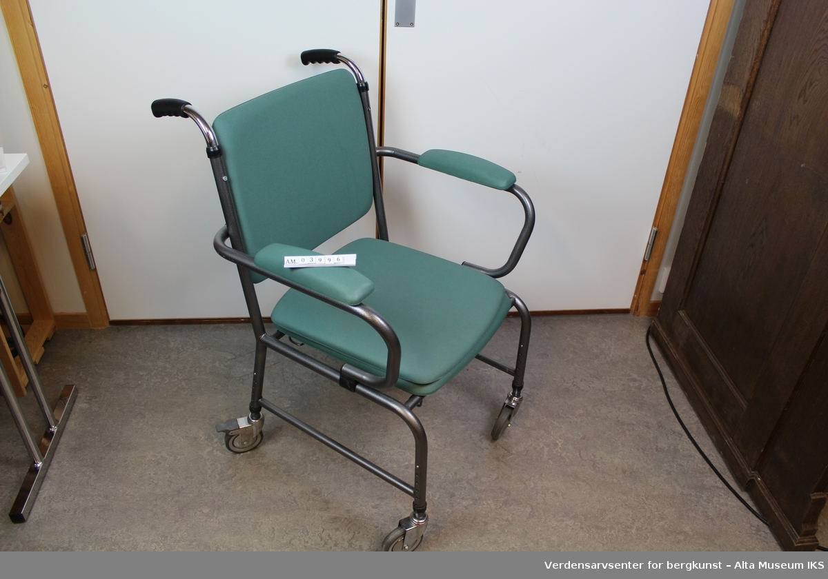 Plastbelagt stålrørstol på hjul. Med avtakbart sete over et dosete og dobøtte. Setet, ryggen og armlenene har puter med et grønnt trekk. Har håndtak for å trille stolen.