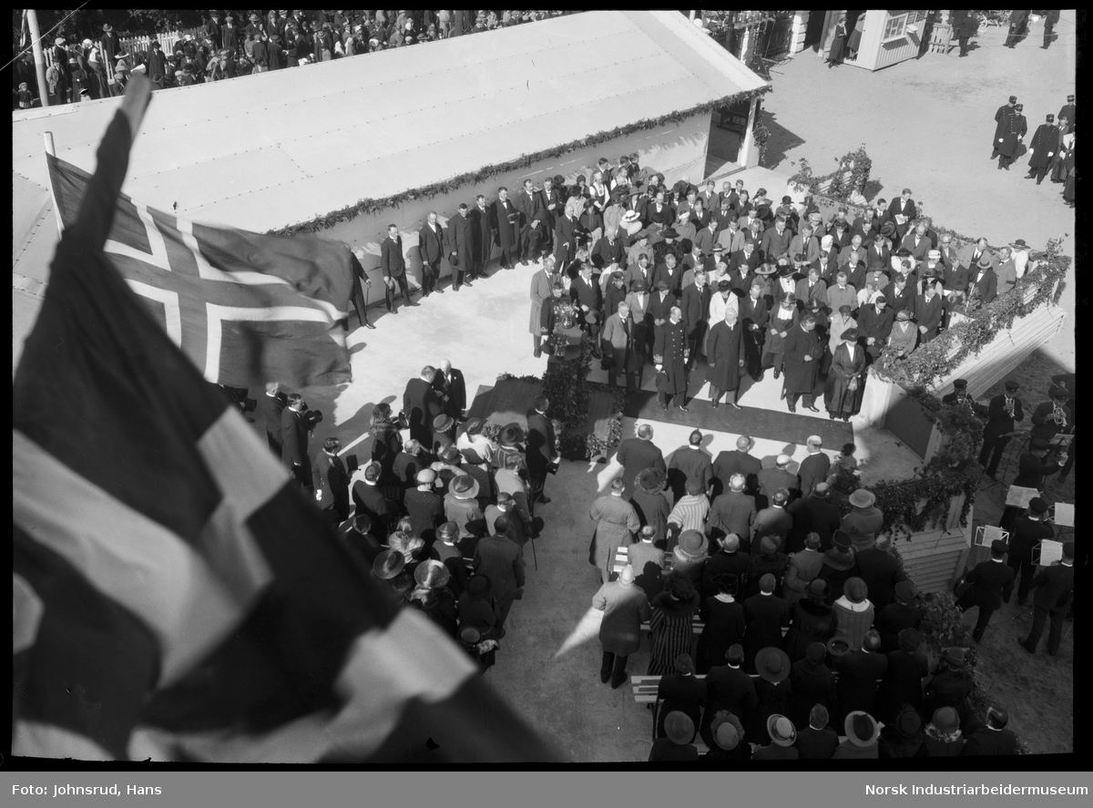 Åpning av Fylkesutstillingen 1922 med besøk av Kong Haakon VII. HM kongen står på rød løper. Korps spiller, jenter i uniform langs veien. Folkemengde rundt hovedarena for åpningsseremonien.