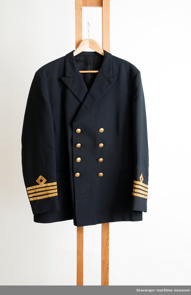 Kapteinsuniform- jakke og bukse i mørk blå ull. Jakken er dobbeltknappet med til sammen 8 knapper i støpt messing med anker. Nedre del av ermer, 4 rader med metallbånd.