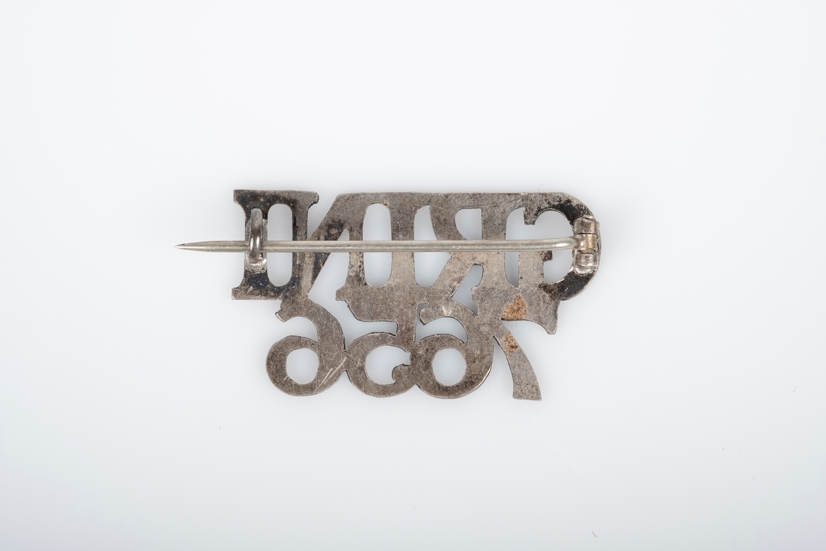 En nål i metall med teksten "Grini 7656". Nål bak som festes i en krok.