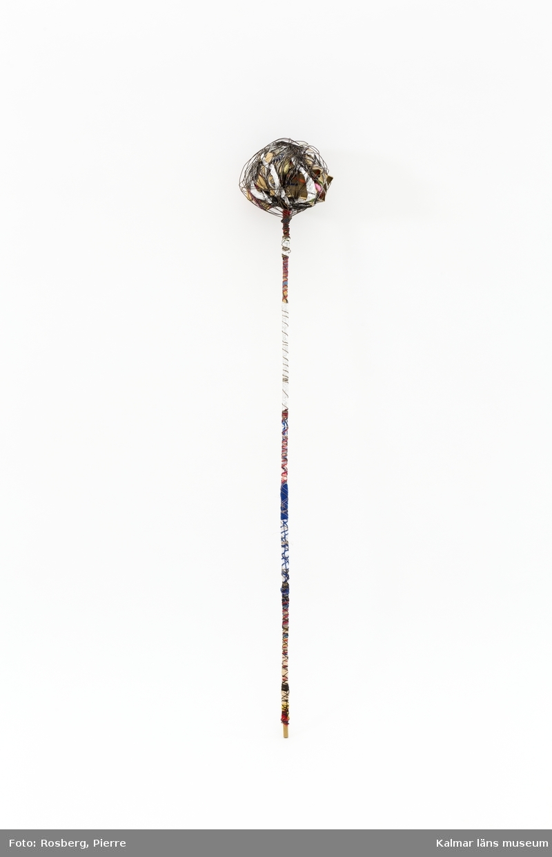 En stav av bambu lindad med band och garn. Högst upp ett klot av tändsticksasklådor som lindats ihop med metall- och sytråd.