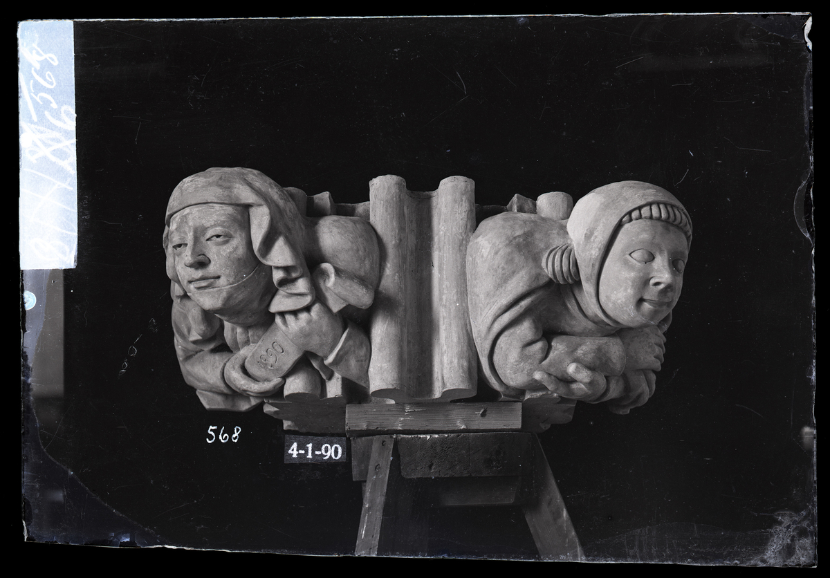 Modell til anfangerfigur i hvelvet i koret i Nidarosdomen. Modellen er av gips. To figurer, en kvinne til venstre som holder ett bånd med årstallet 1890 skrevet på, og en mann med stram lue. Skulptør ukjent.