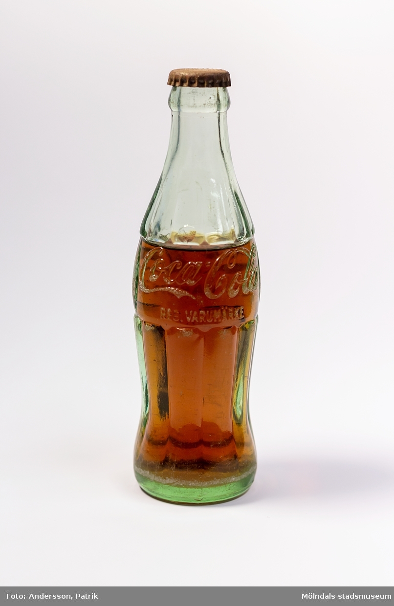 Coca-Cola i glasflaska från i mitten/slutet av 1950-talet. Flaskan är tillverkad i grönaktigt glas. Loggan "Coca-Cola REG. VARUMÄRKE" är inristat i glaset.
Glasflaskans form är inspirerad av kakaobönan. Det geniala med flaskan är dess greppvänliga form med dess särpräglade utseende som gör att man känner igen den även om den greppas i totalt mörker. Glaset splittrades heller inte om flaskan tappades i marken. I början tillverkades flaskorna i färgerna blått, grönt och glasklart glas.

Glasingenjören Alexander Samuelson som formgav flaskan har koppling till Sverige. Han föddes nämligen i Kungälv den 4 januari 1862. Under sina ungdomsår arbetade han på Surte glasbruk. Vid 21 års ålder flyttade han med sin storebror till USA, där han fortsatte att arbeta på olika glasbruk och blev så småingom chef. I början av 1910-talet anordnades en tävling för att designa en flaska till Coca-Colan. Tävlingen vann Alexander och hans glasbruk. 1915 tog Alexander Samuelson patent på denna  geniala utformning av Coca-Colaflaskan, en av världens mest välkända varuförpackningar.

Eftersom livsmedelslagen i Sverige hade förbud mot koffein och fosforsyra, kunde inte Coca-Colan börja säljas i Sverige förrän 1953 då förbudet upphäves. Det var då Pripps bryggerier som tog hem lincensen för att börja tillverkningen av den amerikanska drycken i Göteborg, Malmö och Sundbyberg. 
Coca-Colan hade först svårt att konkurrera med storheterna som sockerdricka, Champis, Festis och Zingo. Det var först i slutet av 1970-talet som försäljningen ökade ordentligt.