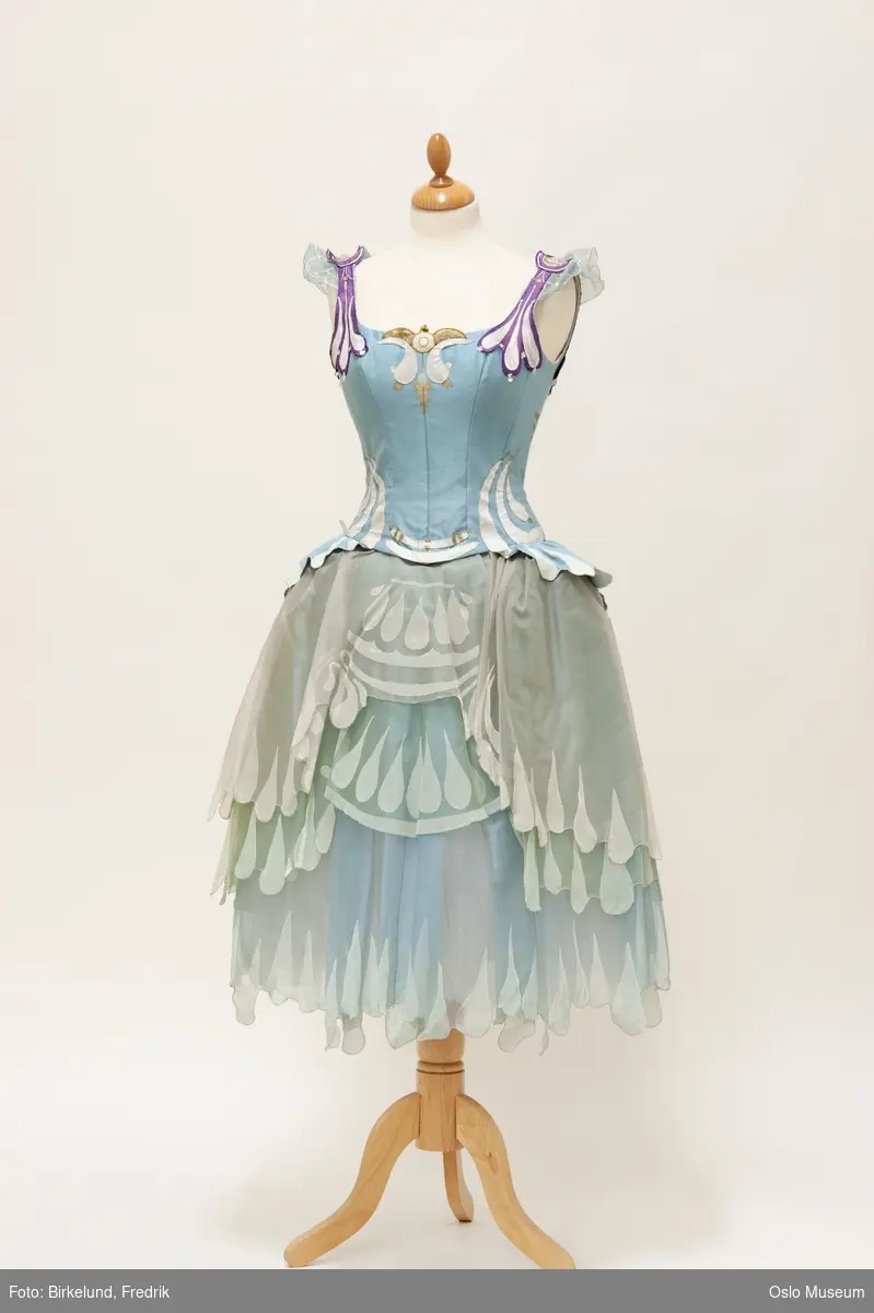 Ballettkjole med lys blå overdel, applikert med hvitt, fiolett og gull. Skjørt i lys blå/ grå organsa, malt med hvitt.