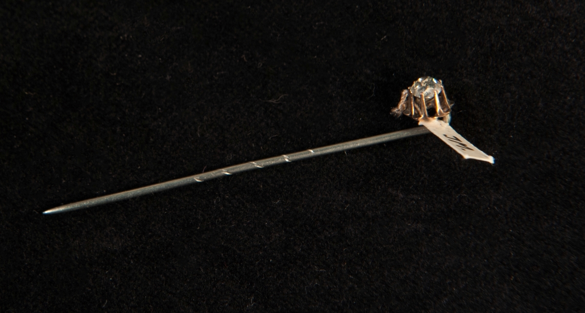 Kråsnål av silver med infattad klar glassten. En svårtydd stämpel finns längst upp på nålen.
