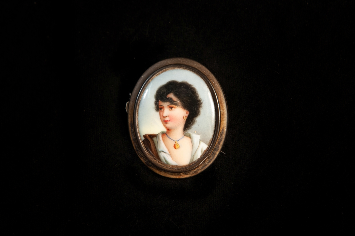 En oval brosch i gråbrunt horn med ett målat porträtt av ung, mörkhårig flicka mot ljus blå bakgrund. Fästnål av mässing. Miniatyr.