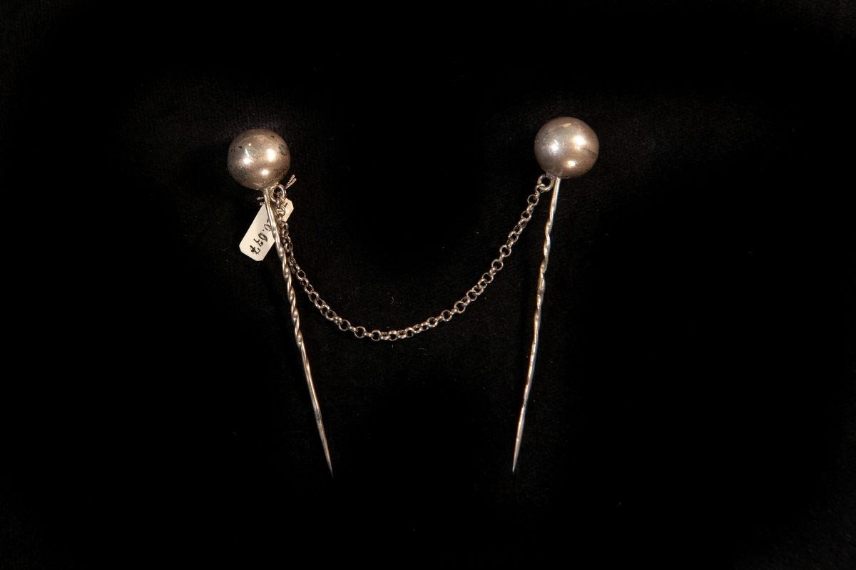 Schalnålar, 2 st (ett par) av silver(?) i form av kula på lång nål förenade med en kedja.