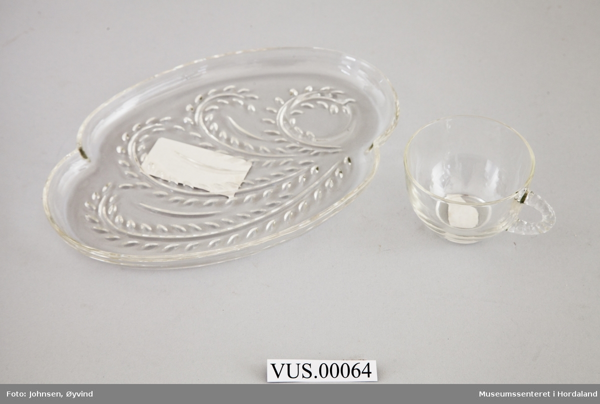 Kopp og tilhørende tallerken i formpresset glass. Tallerkenen er avlang med stråmønster, og har fordypning til koppen. Koppen er glatt med taumønstret håndtak.