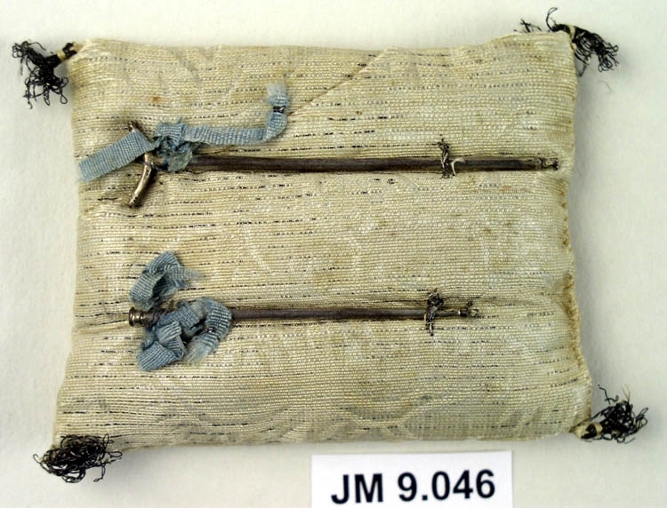 Käpp och krycka av silver monterat på potpouripåse (återanvänd textil med söm på både fram- och baksidan) med små tofsar i hörnen. Små blå rosetter är fästa vid käppen och kryckan.