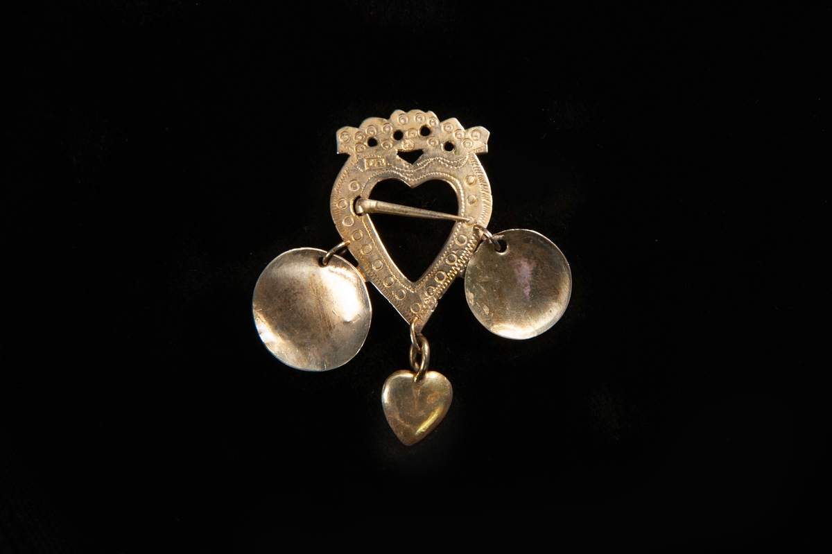 Ett hjärtformat spänne av förgyllt silver. Graverad dekor av punkter m.m. Två skålar och ett hjärta av silver hänger från spännet. Rester efter gravyr på en av skålarna visar att återanvänt material har använts.