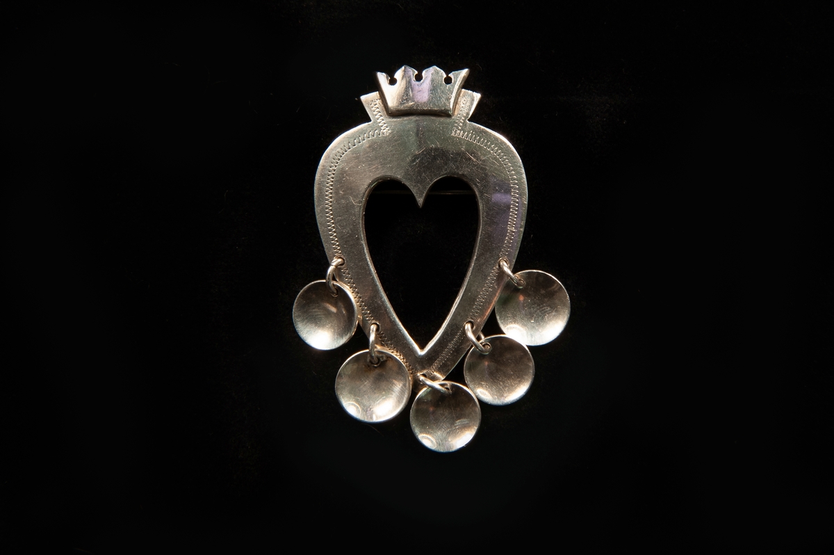 Dräktspänne av silverplåt, hjärtformat med fem kläppen hängande nedtill. Hjärtat har punsad prickdekor och kröns med krona. Baksidan försedd med nål.