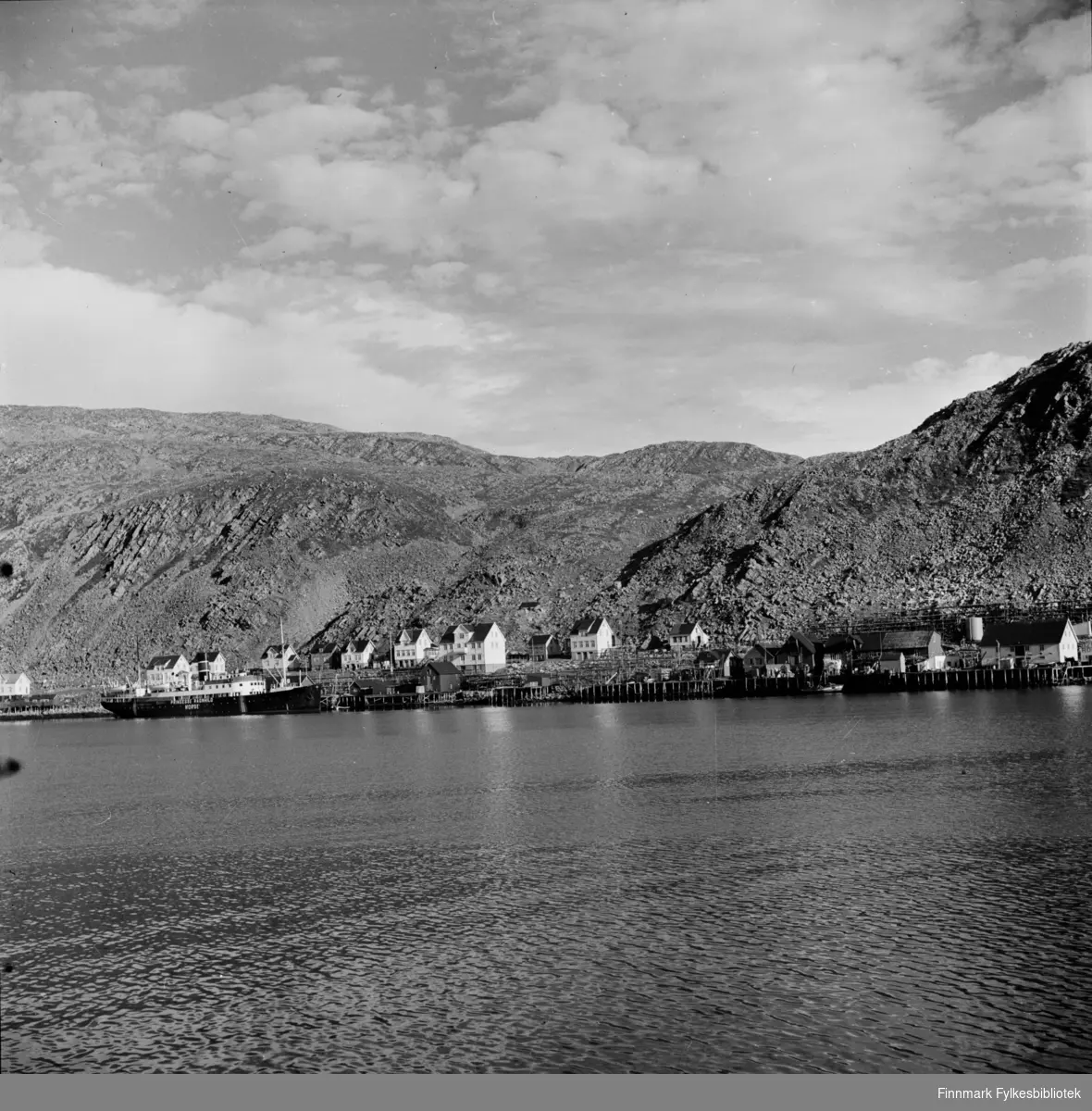 Kjøllefjord 1940 fotografert over fjorden med hurtigruta som ligger til kai ved Horst-ekspedisjonen. Liv Horst, datter av butikkeier og hurtigruteekspeditør Horst, kan huske fotografen Elisabeth Meyer som gikk rundt med flere fotoapparater i Kjøllefjord. Her ligger også fiskebrukene som perler på en snor.
