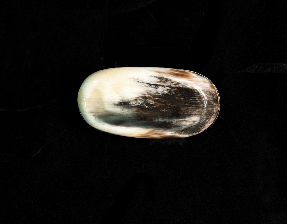 En oval snusdosa av svartvit kohorn med lock m.m. i nysilver.