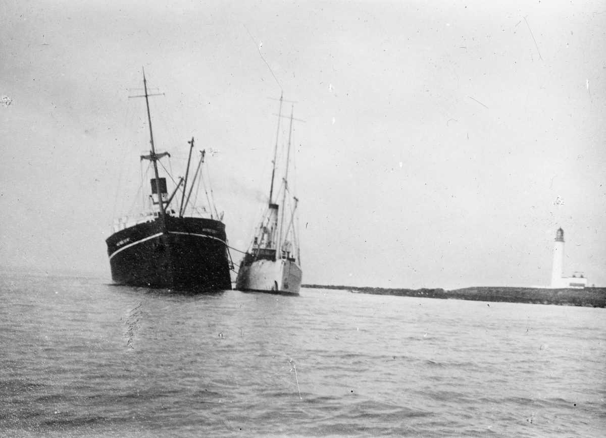 To dampskip i kystlandskap, til høyre i motivet et fyrtårn. Fartøyet til venstre i motivet har slagside.