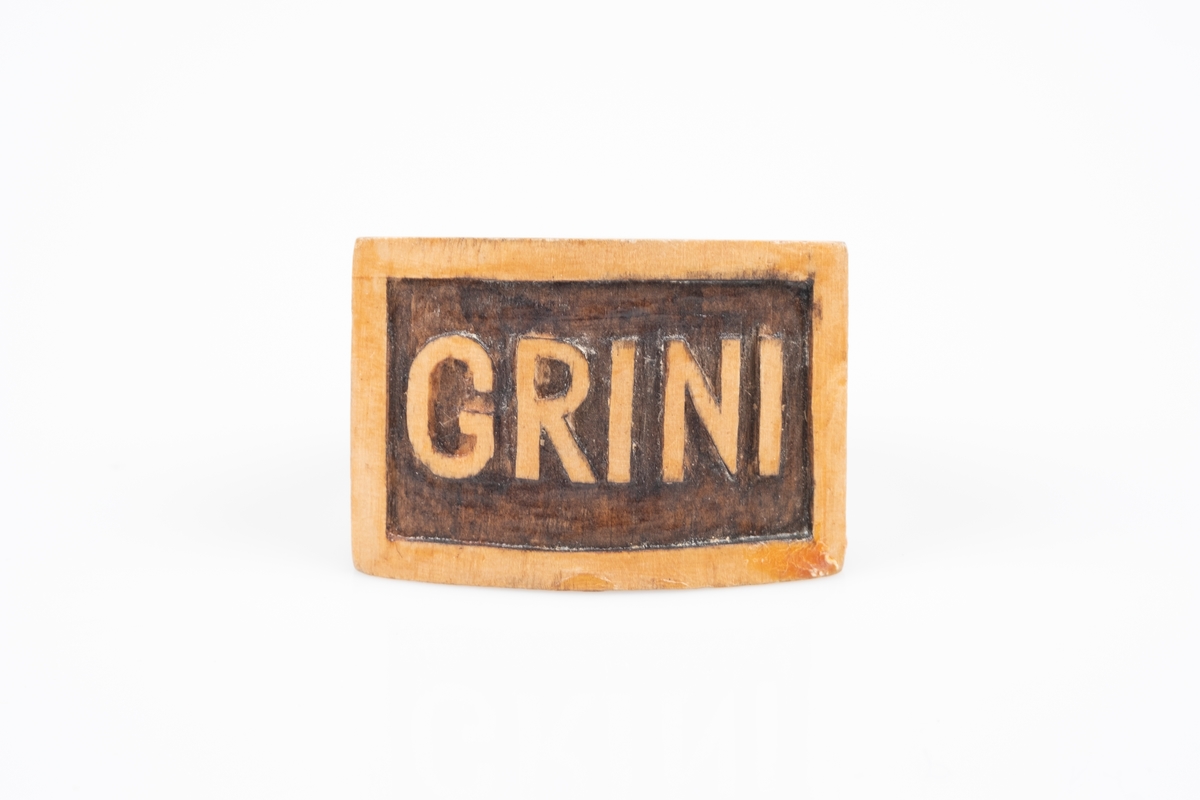 En serviettring i tre. Framsiden er flat og firkantet med utskjæring av ordet "Grini".