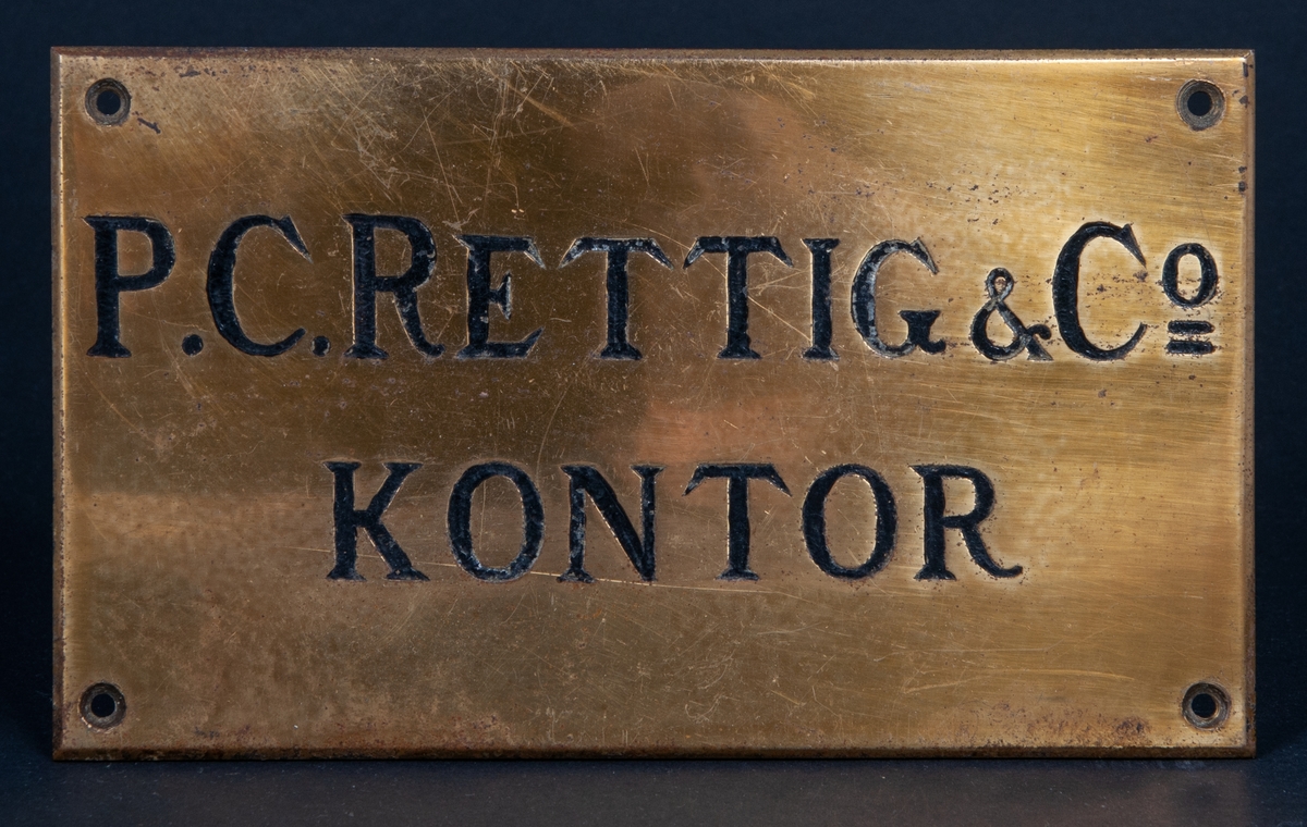 Namnskylt av mässing. Rektangulär svartmålad inskrift: "P.C. RETTIG & Co KONTOR"