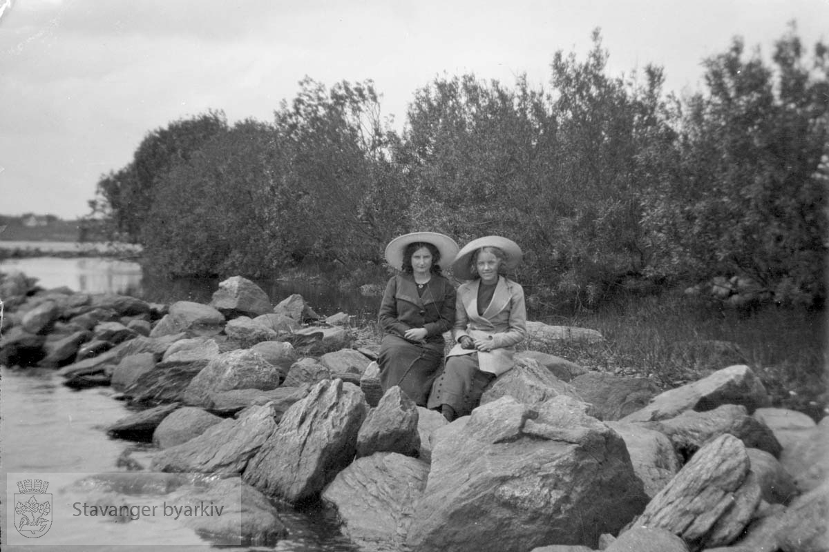 To kvinner med hatt på steiner langs vannkanten