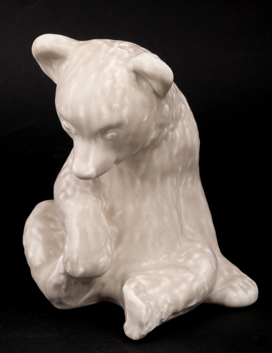 Figurinen Brumsi i gråvit glasyr designad av Lillemor Mannerheim, 1955. Björnen är avbildad i sittande ställning med nedböjt huvud, och lyfter sin högra fot med höger ram.