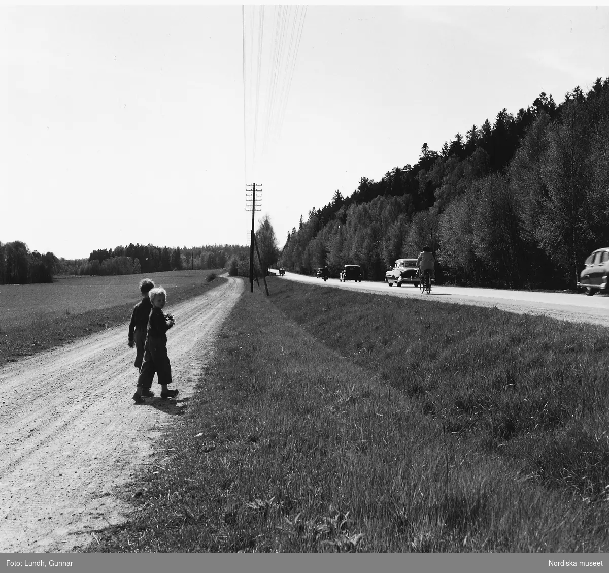 Två barn går på en grusväg  bredvid en större asfalterad väg som trafikeras av bilar och en cyklist.