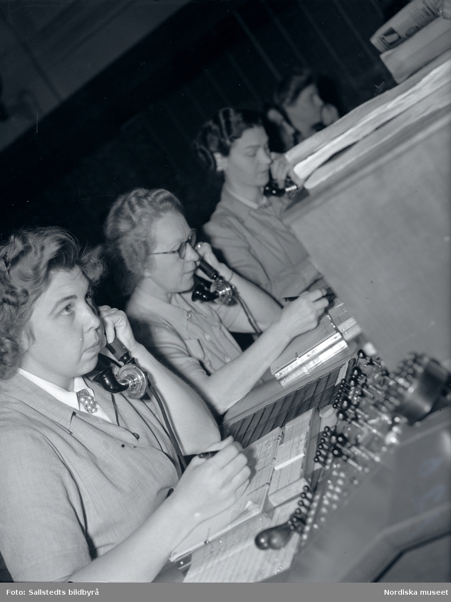 Telefonister på Stockholms telefonstation år 1946. En rad med kvinnor sitter vid ett växelbord och lyssnar i telefonlur och antecknar i anteckningsblock.
