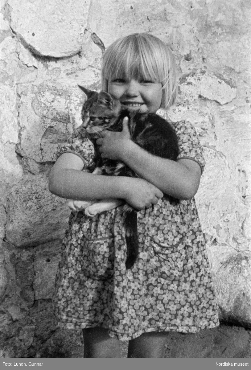 Porträtt av två flickor som står vid en husvägg och håller en katt i famnen, porträtt av en flicka som håller en katt i famnen, fotograferat vid Kullen (Kullaberg), Strandbaden, Tallbacken.