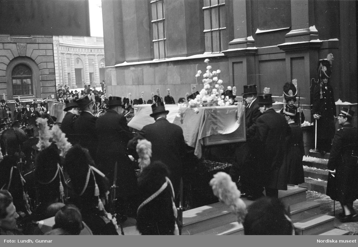 Troligen begravningen av Prins Gustaf Adolf, en kista bärs ut från Storkyrkan, kungafamiljen går ut från Storkyrkan, militärer i uniform står i givakt, kistan står på en katafalk som dras av hästar, en folksamling står längs vägen när begravningsföljet passerar.