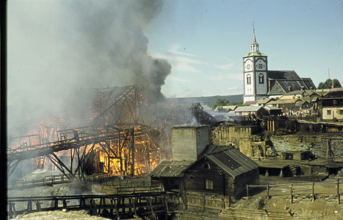 Brann i smeltehytta på Røros, lørdag 19. juli 1975, sett fra østsida av elva. Foran den brennende smeltehytta sees øvre maskinhuset og sleggbanen som går over til østsida av elva